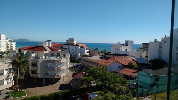 A coastal urban area with white and red-roofed buildings is visible, surrounded by greenery. In the distance, the calm sea is present under a clear blue sky. The setting suggests a quiet, sunny seaside neighborhood.