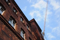 A tall brick building is captured from a low angle, showcasing its intricate window designs and the texture of the brickwork. A construction crane is visible in the background under a partly cloudy sky, indicating possible construction or renovation activity.