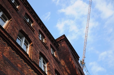 A tall brick building is captured from a low angle, showcasing its intricate window designs and the texture of the brickwork. A construction crane is visible in the background under a partly cloudy sky, indicating possible construction or renovation activity.