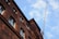 A tall brick building is captured from a low angle, showcasing its intricate window designs and the texture of the brickwork. A construction crane is visible in the background under a partly cloudy sky, indicating possible construction or renovation activity.