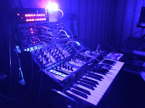 A dimly lit room features an array of electronic music equipment, including a synthesizer and a complex system of interconnected cables. A glowing blue and purple light bathes the equipment, while several control panels with knobs and buttons are visible.