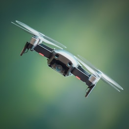 A drone is hovering in the air against a blurred green background. The drone is compact with visible propellers spinning rapidly. Its camera is prominently displayed on the front, indicating that it is likely used for aerial photography or videography.