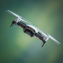 A drone is hovering in the air against a blurred green background. The drone is compact with visible propellers spinning rapidly. Its camera is prominently displayed on the front, indicating that it is likely used for aerial photography or videography.