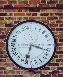 A large round clock with Roman numerals is mounted on a brick wall. The clock face has 'Shepherd Patentee' and 'Calvanic Magnetic Clock' written on it.