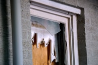 A close-up view of a broken window, with shattered glass pieces and torn paper or cardboard covering the opening. The window frame appears worn and is set against a brick wall.