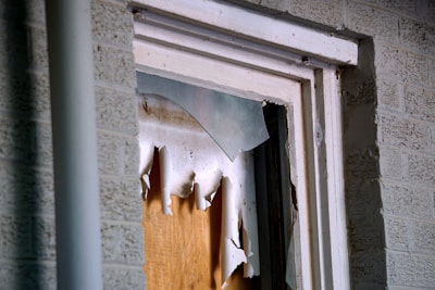 A close-up view of a broken window, with shattered glass pieces and torn paper or cardboard covering the opening. The window frame appears worn and is set against a brick wall.