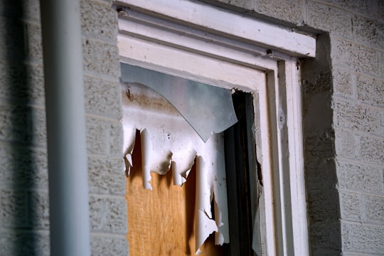 A close-up view of a broken window, with shattered glass pieces and torn paper or cardboard covering the opening. The window frame appears worn and is set against a brick wall.