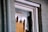 A close-up view of a broken window, with shattered glass pieces and torn paper or cardboard covering the opening. The window frame appears worn and is set against a brick wall.