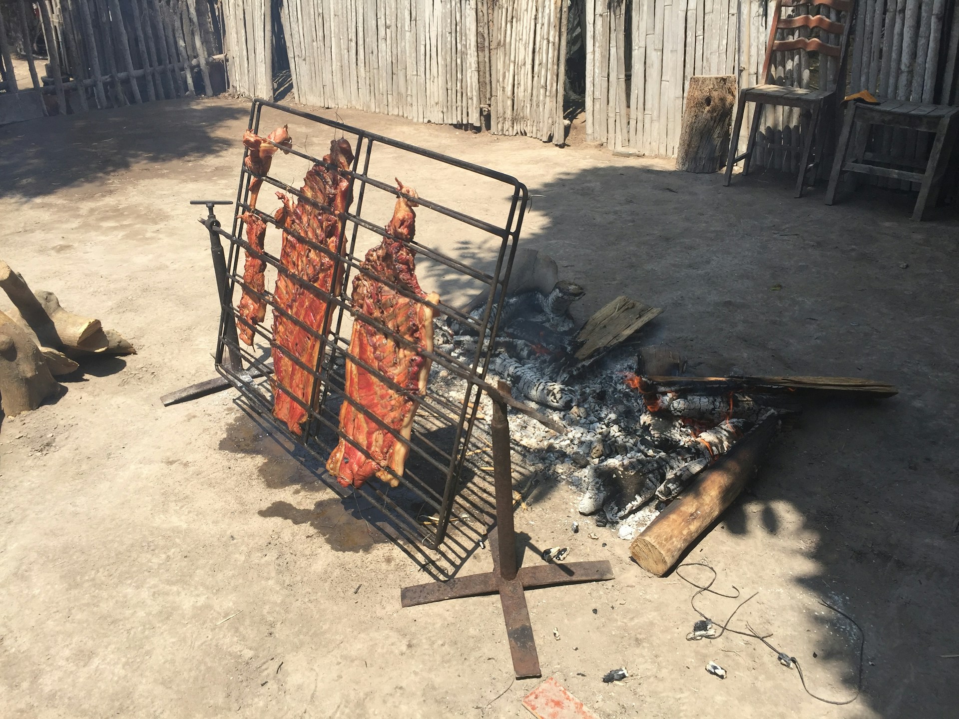 Several large pieces of meat are being cooked on a vertical metal grill over an open fire. The scene is outdoors, possibly in a rustic or rural setting, with rough wooden fencing and simple chairs visible in the background.