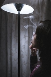 A person is exhaling smoke while sitting next to a lit floor lamp. The room appears dimly lit, with a floral patterned curtain in the background. The scene has a contemplative and quiet atmosphere.