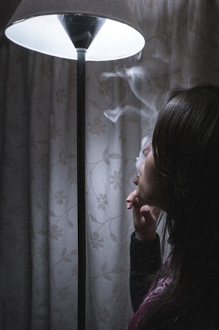 A person is exhaling smoke while sitting next to a lit floor lamp. The room appears dimly lit, with a floral patterned curtain in the background. The scene has a contemplative and quiet atmosphere.