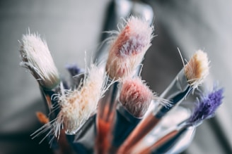 Several close-up paintbrushes with frayed bristles, each showing a variety of pastel and earthy tones. The bristles appear soft and fluffy, suggesting frequent use. The background is softly blurred, emphasizing the focus on the textured bristles of the brushes.