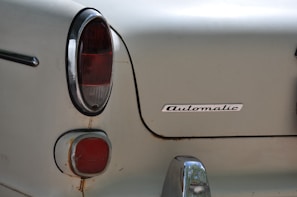 The image captures the rear section of an old car featuring a classic, rounded tail light design. The word 'Automatic' is prominently displayed on the metal body, which appears slightly weathered with some rust spots. A small section of a chrome bumper is visible at the lower edge.