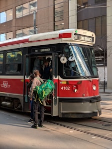 A red and black streetcar with the route number 501 Long Branch is stopped on a city street. Several people, including one carrying a large green backpack, are boarding the vehicle. It appears to be a busy urban area with tall buildings in the background.