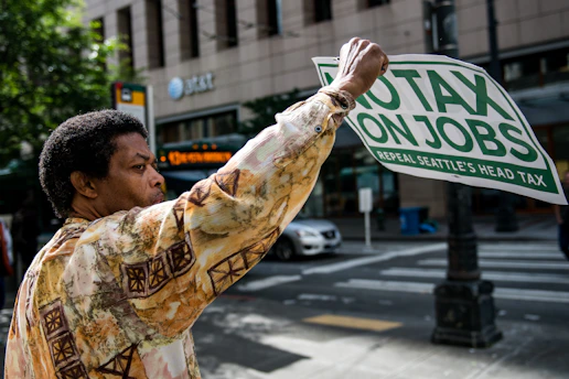 A person is holding a sign that reads 'No Tax On Jobs, Repeal Seattle's Head Tax' while standing on a city street. They wear a patterned shirt, and in the background, there are trees, a building with AT&T signage, and a passing car.