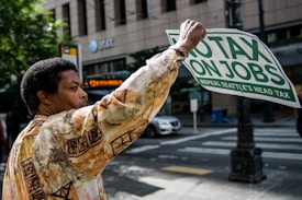 A person is holding a sign that reads 'No Tax On Jobs, Repeal Seattle's Head Tax' while standing on a city street. They wear a patterned shirt, and in the background, there are trees, a building with AT&T signage, and a passing car.