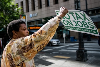 A person is holding a sign that reads 'No Tax On Jobs, Repeal Seattle's Head Tax' while standing on a city street. They wear a patterned shirt, and in the background, there are trees, a building with AT&T signage, and a passing car.