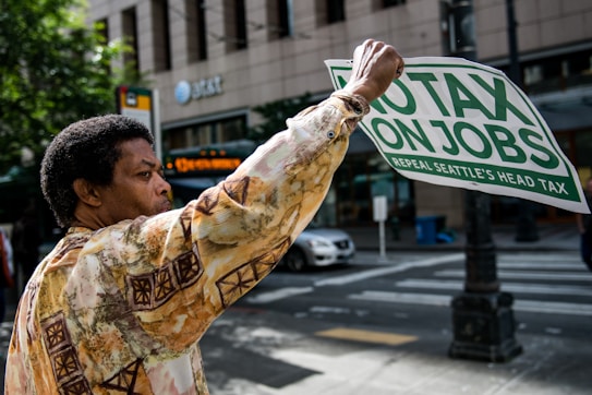 A person is holding a sign that reads 'No Tax On Jobs, Repeal Seattle's Head Tax' while standing on a city street. They wear a patterned shirt, and in the background, there are trees, a building with AT&T signage, and a passing car.