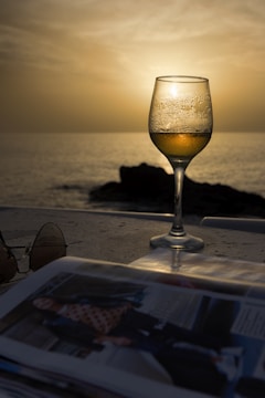 A wine glass filled with a chilled beverage is placed on a table with a scenic beach sunset in the background. On the table, there are also sunglasses and an open magazine. The sea is calm and the sky is golden with the setting sun creating a peaceful ambiance.
