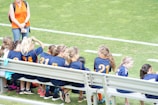 A group of young girls in blue and orange sports jerseys is seated on a metal bench beside a soccer field. The grass is bright green, indicating a well-maintained sports ground. A woman in an orange top and jeans stands nearby, possibly a coach or a parent.