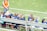 A group of young girls in blue and orange sports jerseys is seated on a metal bench beside a soccer field. The grass is bright green, indicating a well-maintained sports ground. A woman in an orange top and jeans stands nearby, possibly a coach or a parent.