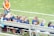 A group of young girls in blue and orange sports jerseys is seated on a metal bench beside a soccer field. The grass is bright green, indicating a well-maintained sports ground. A woman in an orange top and jeans stands nearby, possibly a coach or a parent.