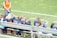 A group of young girls in blue and orange sports jerseys is seated on a metal bench beside a soccer field. The grass is bright green, indicating a well-maintained sports ground. A woman in an orange top and jeans stands nearby, possibly a coach or a parent.