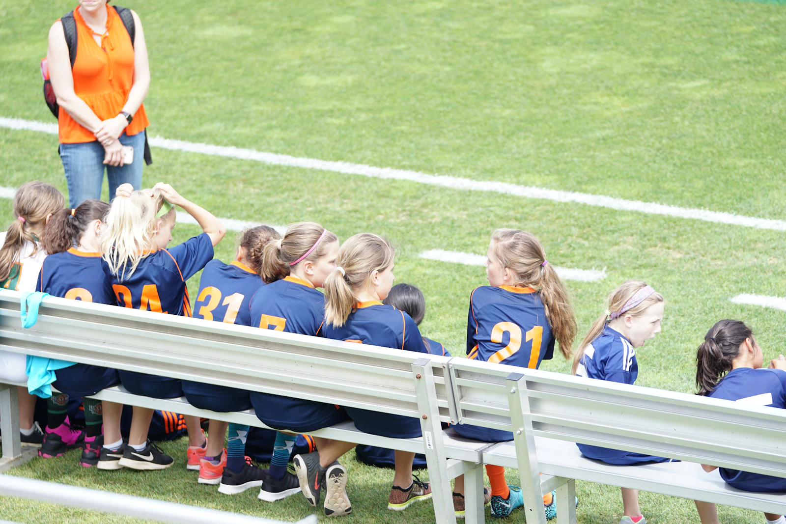 A youth girls' soccer team in uniform sitting together on a bench beside a green field.