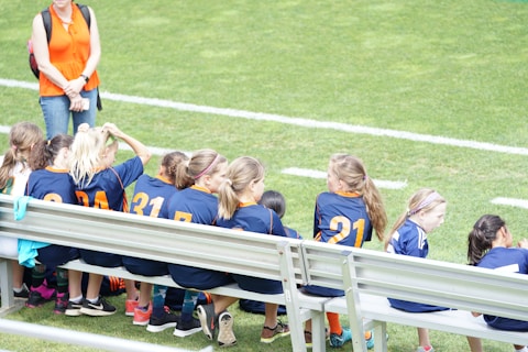 A group of young girls in blue and orange sports jerseys is seated on a metal bench beside a soccer field. The grass is bright green, indicating a well-maintained sports ground. A woman in an orange top and jeans stands nearby, possibly a coach or a parent.