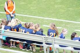 A group of young girls in blue and orange sports jerseys is seated on a metal bench beside a soccer field. The grass is bright green, indicating a well-maintained sports ground. A woman in an orange top and jeans stands nearby, possibly a coach or a parent.