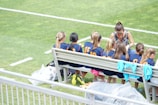 A group of young athletes in matching blue uniforms with orange numbers sit on a bench beside a coach on a grassy field. The coach appears to be giving instructions or advice. The scene is bright and sunny, with some sports jerseys draped over the bench.