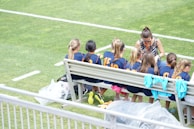A group of young athletes in matching blue uniforms with orange numbers sit on a bench beside a coach on a grassy field. The coach appears to be giving instructions or advice. The scene is bright and sunny, with some sports jerseys draped over the bench.