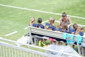 A group of young athletes in matching blue uniforms with orange numbers sit on a bench beside a coach on a grassy field. The coach appears to be giving instructions or advice. The scene is bright and sunny, with some sports jerseys draped over the bench.