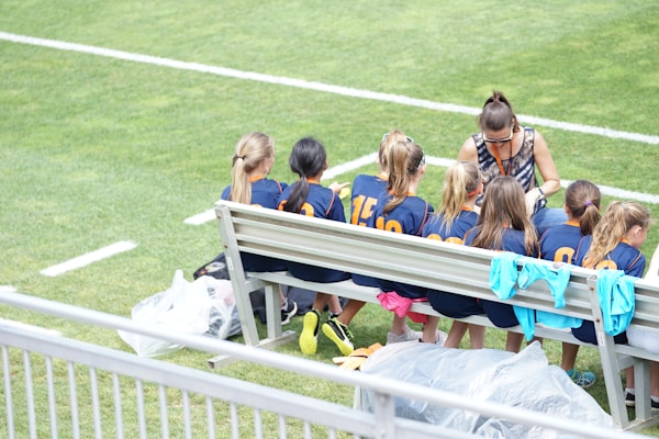 A group of young athletes in matching blue uniforms with orange numbers sit on a bench beside a coach on a grassy field. The coach appears to be giving instructions or advice. The scene is bright and sunny, with some sports jerseys draped over the bench.