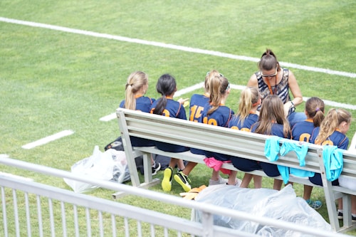 A group of young athletes in matching blue uniforms with orange numbers sit on a bench beside a coach on a grassy field. The coach appears to be giving instructions or advice. The scene is bright and sunny, with some sports jerseys draped over the bench.