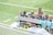 A group of young female athletes wearing blue jerseys with numbers sit on a bench on a grassy field. An adult female, possibly a coach, is interacting with them, holding a piece of paper. The area is bordered by a white fence, and clothes are draped over the back of the bench.