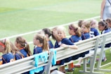 A group of young girls wearing blue sports jerseys are seated on a bench by the side of a grassy field. They appear to be part of a soccer team, with some shirts draped over the bench. The girls have various hairstyles, including ponytails, and one girl in the center is making a funny face.