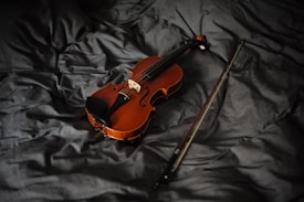 A violin rests on a rumpled, dark fabric surface, with its bow placed nearby. The lighting casts a warm glow on the polished wooden body of the violin, highlighting its fine craftsmanship and curves.