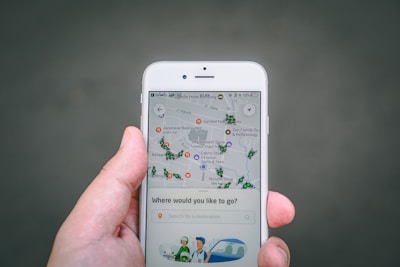 A hand holds a smartphone displaying a map application. The map shows various locations marked with icons. The screen also has a search bar with the prompt 'Where would you like to go?' and an illustration of a driver and a car at the bottom.