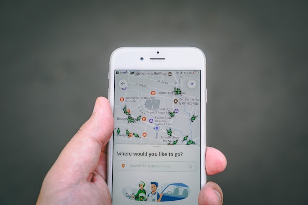 A hand holds a smartphone displaying a map application. The map shows various locations marked with icons. The screen also has a search bar with the prompt 'Where would you like to go?' and an illustration of a driver and a car at the bottom.