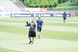 A person in a sports jacket and shorts is juggling a soccer ball on their head on a grassy field. Another person in a t-shirt and shorts walks away in the background. The field is part of a stadium with empty bleachers and advertising banners. Trees and a fence are visible in the background.
