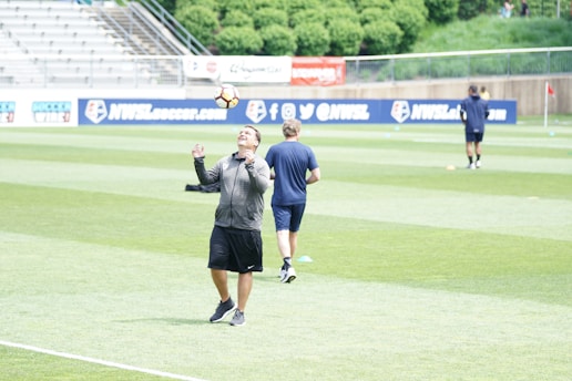 A person in a sports jacket and shorts is juggling a soccer ball on their head on a grassy field. Another person in a t-shirt and shorts walks away in the background. The field is part of a stadium with empty bleachers and advertising banners. Trees and a fence are visible in the background.