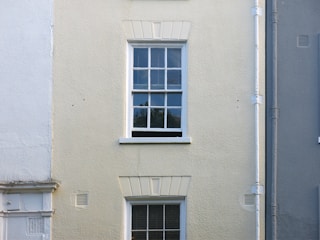 A cream-colored wall features a single, white-framed window with multiple panes. The wall has a rough texture, with a vertical drainage pipe running alongside the window. The lower part of the wall has faint traces of white paint, blending with the cream surface.