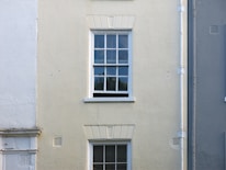A cream-colored wall features a single, white-framed window with multiple panes. The wall has a rough texture, with a vertical drainage pipe running alongside the window. The lower part of the wall has faint traces of white paint, blending with the cream surface.