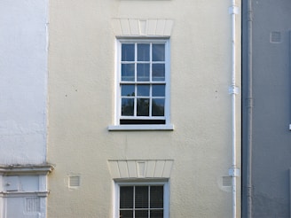 A cream-colored wall features a single, white-framed window with multiple panes. The wall has a rough texture, with a vertical drainage pipe running alongside the window. The lower part of the wall has faint traces of white paint, blending with the cream surface.