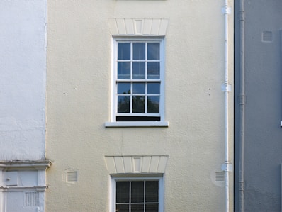 A cream-colored wall features a single, white-framed window with multiple panes. The wall has a rough texture, with a vertical drainage pipe running alongside the window. The lower part of the wall has faint traces of white paint, blending with the cream surface.