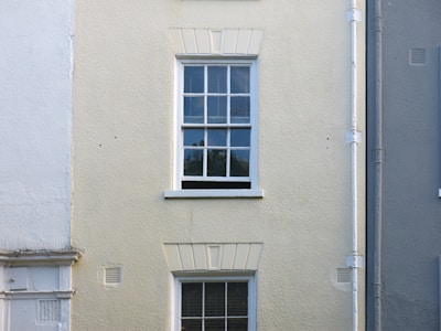 A cream-colored wall features a single, white-framed window with multiple panes. The wall has a rough texture, with a vertical drainage pipe running alongside the window. The lower part of the wall has faint traces of white paint, blending with the cream surface.