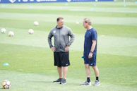 Two people standing on a grass field, possibly a football pitch, surrounded by scattered cones and footballs. Both are dressed in sportswear, engaging in conversation.
