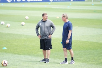 Two people standing on a grass field, possibly a football pitch, surrounded by scattered cones and footballs. Both are dressed in sportswear, engaging in conversation.