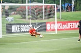 A soccer player wearing a red jersey and orange gloves is crouching on the grass inside the goal area, seemingly in motion to intercept a soccer ball. The goal net and surrounding field are visible in the background. On the left side, part of an advertising board with 'NIKE SOCCER' text is displayed, along with a hashtag referencing a TV network. Spectators and greenery area are visible in the distance.